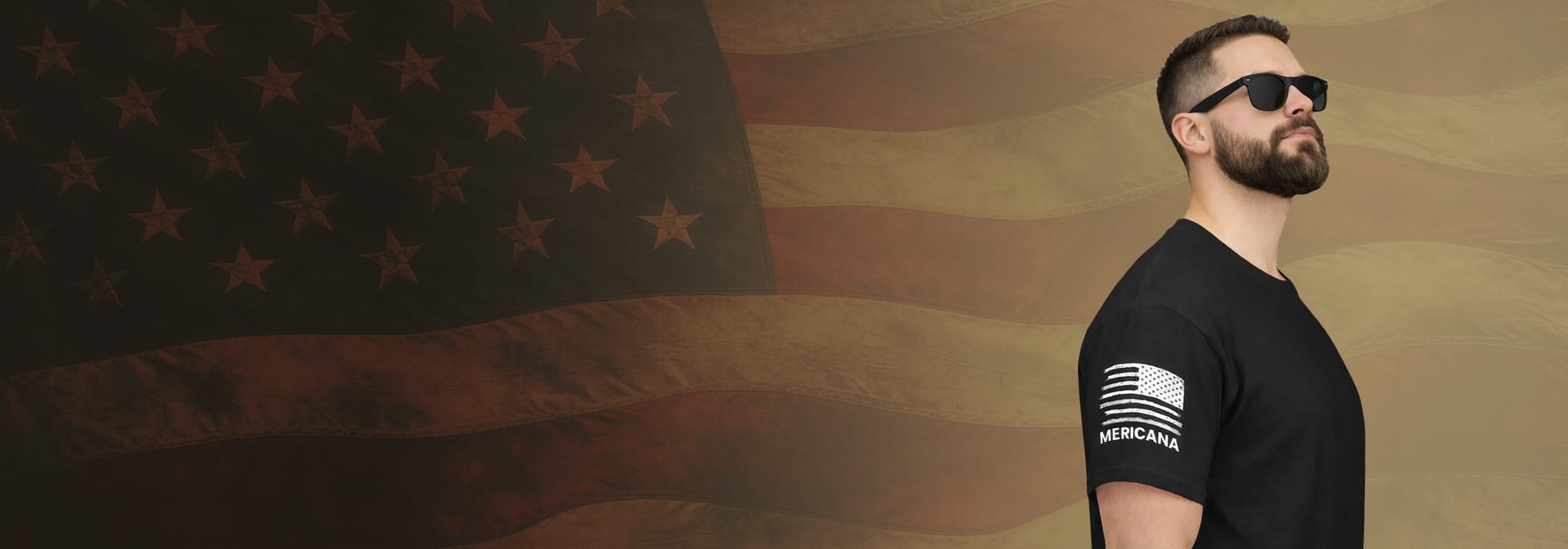 Brunette bearded man with sunglasses wearing black t shirt with Mericana and a white distressed American Flag on the right sleeve, in front of a faded American Flag backdrop