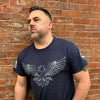 Man standing in front of a brick wall wearing a navy blue shirt with the white outline of an eagle on the front and designs on the sleeves