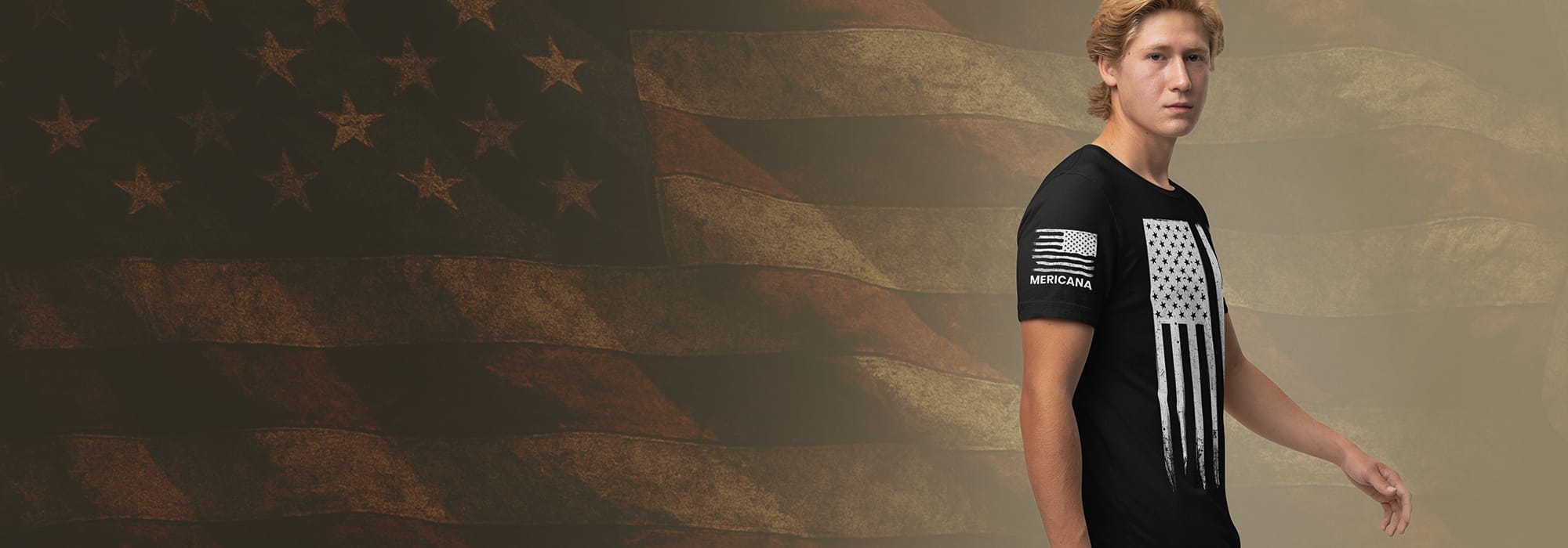 Young blonde man wearing black t shirt with vertical distressed American flag on it, sleeve says Mericana and has a horizontal distressed flag with canton on right