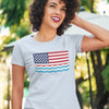 Woman wearing a light blue shirt with an American Flag that has pine trees replacing the stars in the canton and ocean waves below the flag